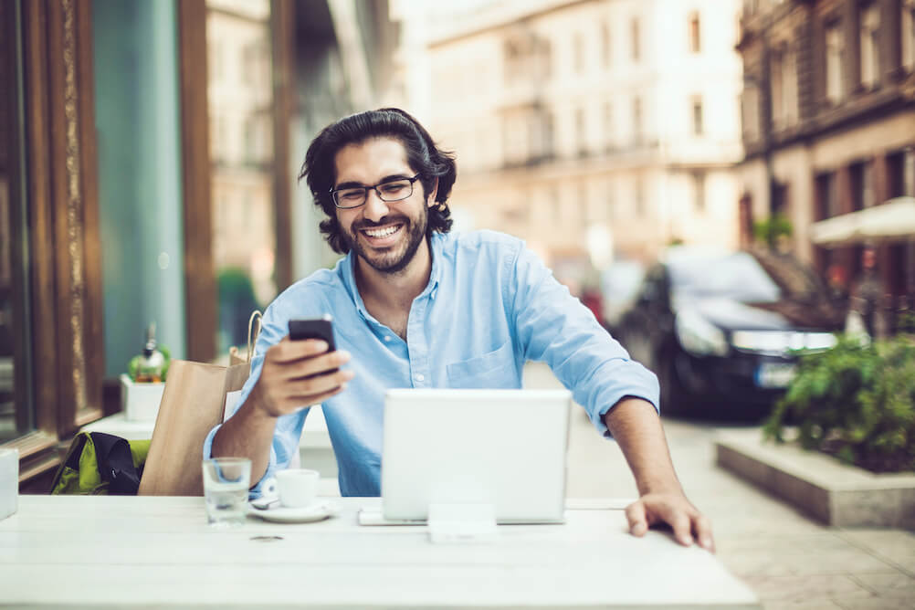 Man at cafe with laptop and looking at phone smiling